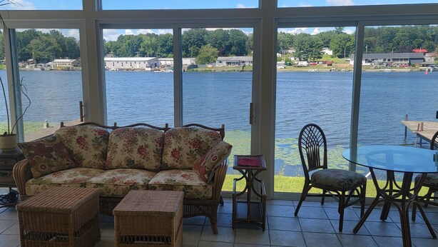 A room with a large window overlooking a lake, a floral sofa, wicker chairs, and a small table. - Seneca Riverfront Spa! Hot tub, Pool + Sauna! Pontoon Boat and Canoe Available! (Baldwinsville)