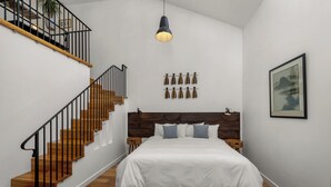 A bedroom with a wooden staircase, a bed with a white bedspread, a hanging light fixture, and a framed picture on the wall.