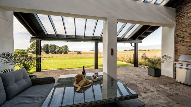 An outdoor patio with a glass table, a sofa, a grill, and a view of the countryside.
