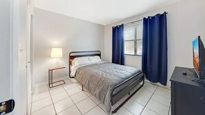 A bedroom with a bed, a bedside table with a lamp, a TV on a stand, and a window with curtains. - Fuseball & Games - 2 BD / 2 BTH Condo Miami! (Miami)