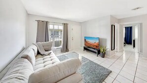 A modern living room with a beige sofa, a flat-screen TV, and a large window with curtains. - Fuseball & Games - 2 BD / 2 BTH Condo Miami! (Miami)