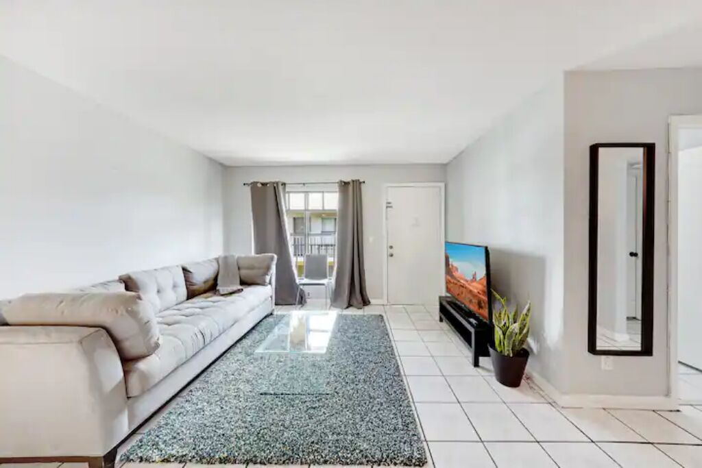 A modern living room with a sofa, a flat-screen TV, a potted plant, and a large mirror.