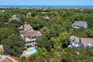An aerial view of a residential area with houses surrounded by dense trees, a swimming pool, and a view of the ocean.
