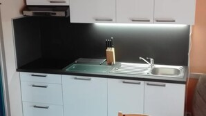 A compact kitchen with white cabinets, a black countertop, and a sink. There is a wooden chair and a table with a patchwork tablecloth.