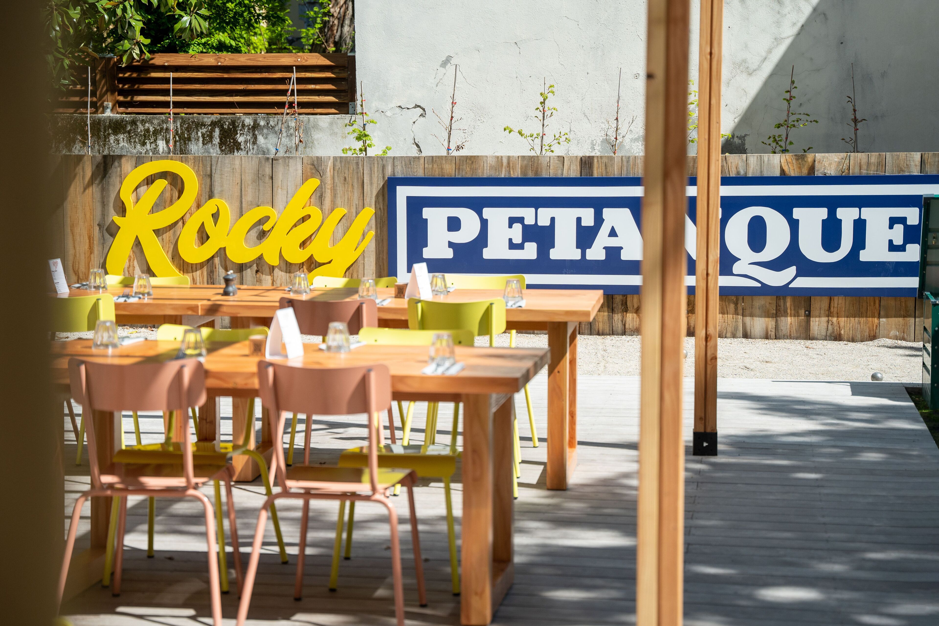 An outdoor seating area with wooden tables and chairs, a large sign reading "Rocky PETAQUE," and a wooden deck.