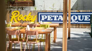 An outdoor seating area with wooden tables and chairs, a large sign reading "Rocky PETAQUE," and a wooden deck.