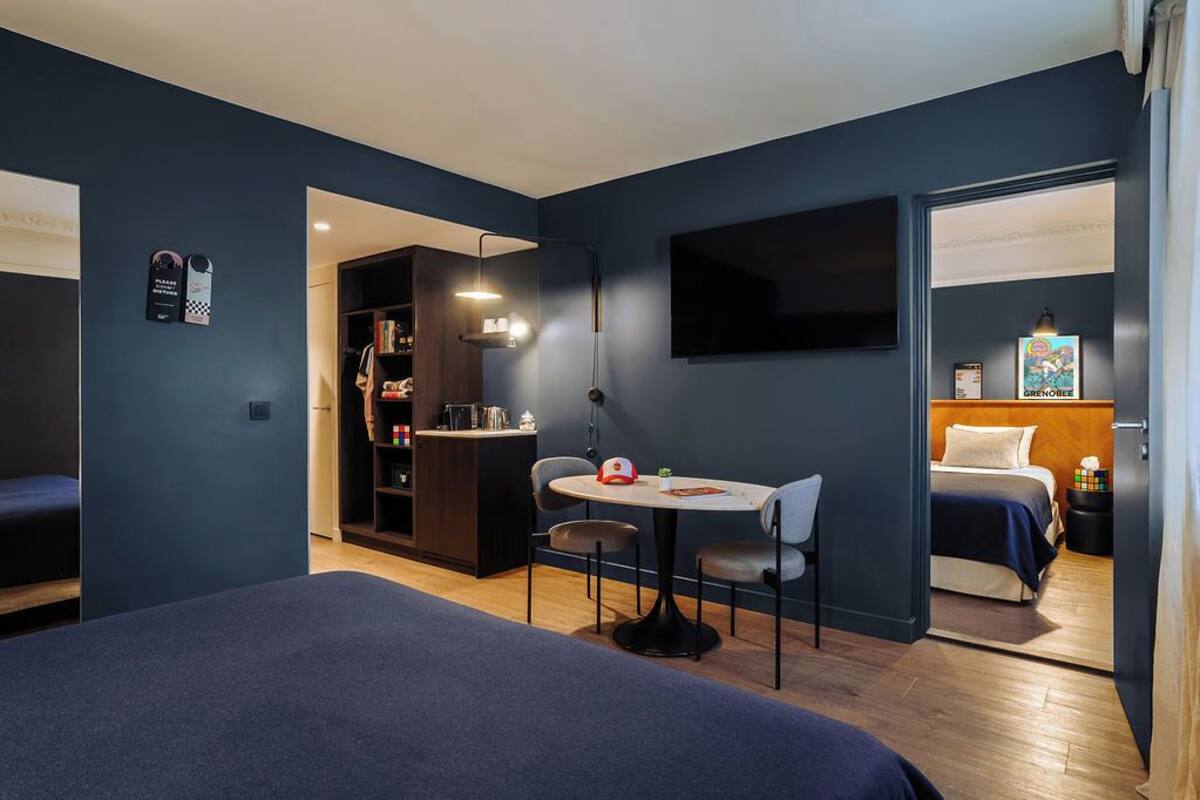 A modern hotel room with a bed, a small dining table, a TV, and a kitchenette.