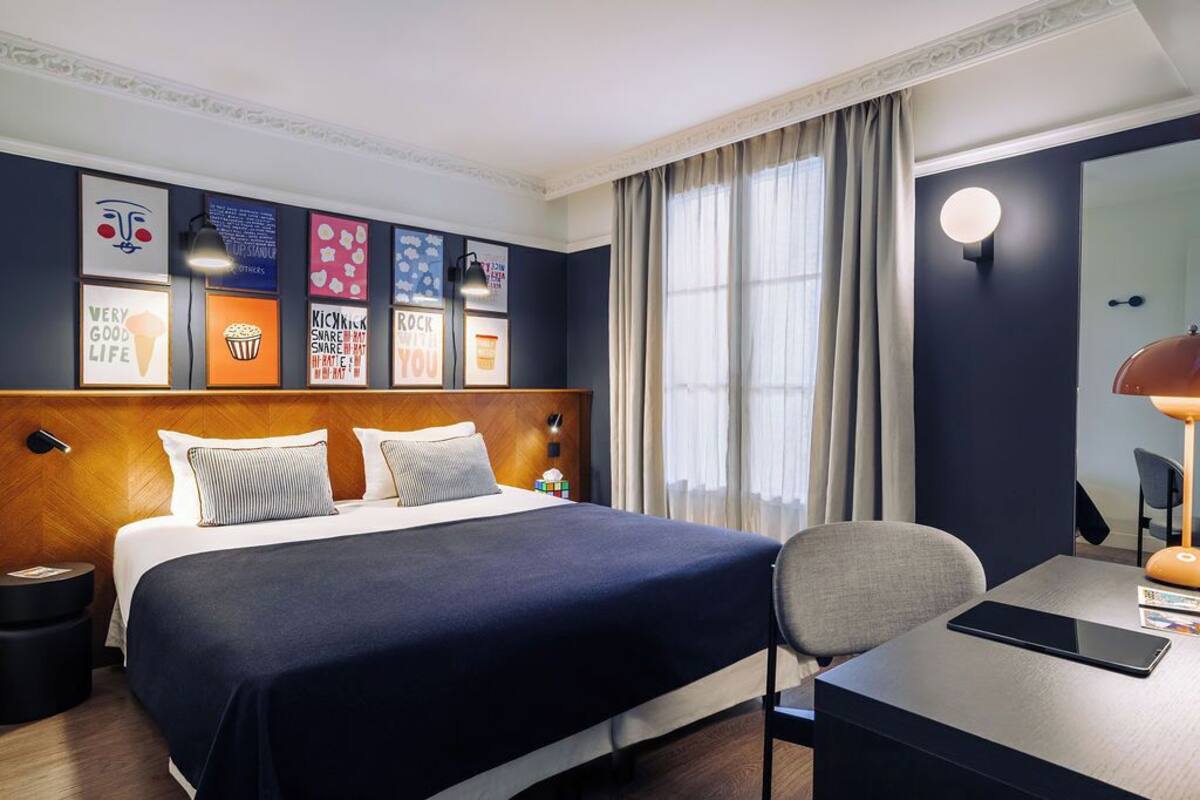 A hotel room with a large bed, a desk with a chair, a round mirror, and framed pictures on the wall.