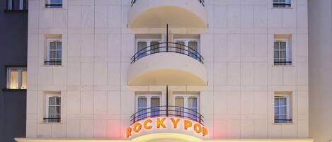 Un hotel de varios pisos con un letrero llamativo que dice "ROCKY POP", una escalera que conduce a la entrada y un área visible de restaurante o bar con señalización.