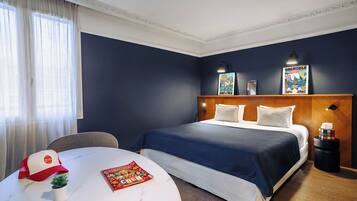 A hotel room with a large bed, a round table with a magazine, a small plant, a red and white cap, and a Rubik's cube on a side table.