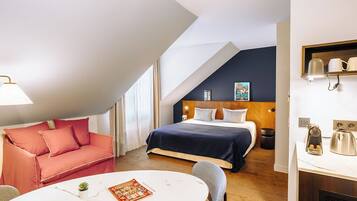 A modern hotel room with a bed, a sofa, a dining table, and a kitchenette.