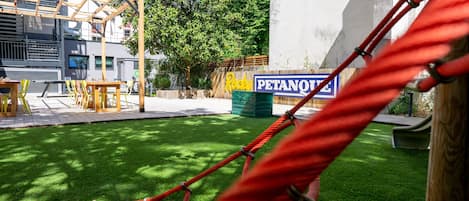 Un parque infantil con un columpio de cuerda, una mesa de picnic y sillas. Se ve un letrero que dice 'Rocky Petanque' al fondo.