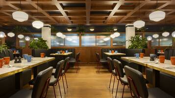 Een restaurant met houten balken in het plafond, ronde hanglampen en zwarte stoelen met rode poten. Tafels hebben oranje en blauwe kopjes en schotels.