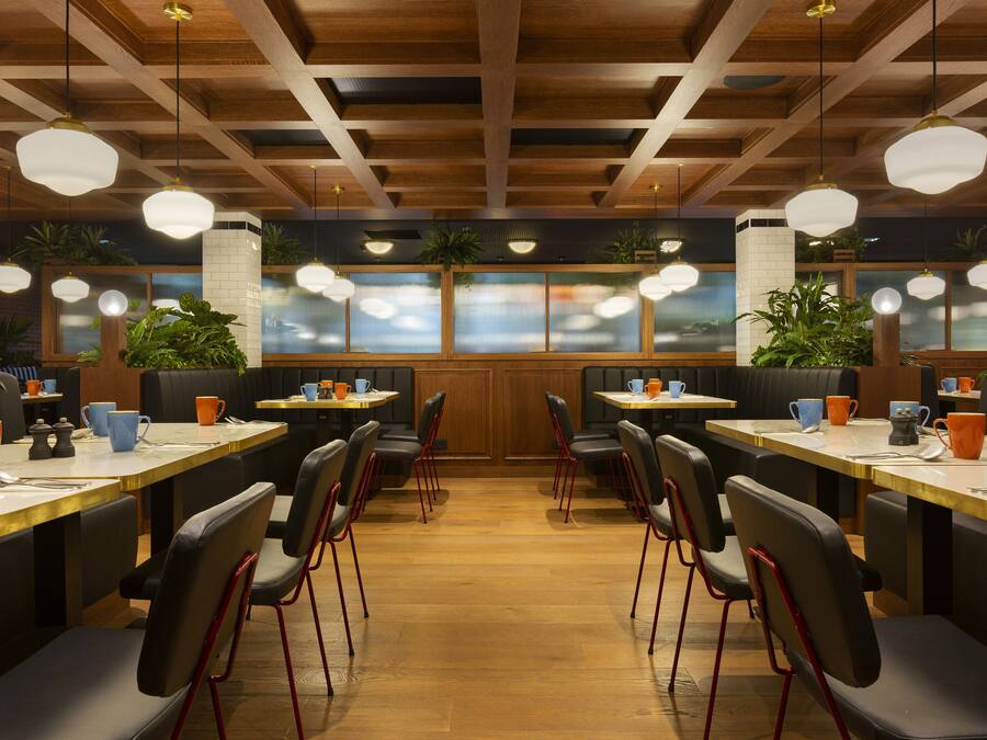 Een restaurant met houten balken in het plafond, ronde hanglampen en zwarte stoelen met rode poten. Tafels hebben oranje en blauwe kopjes en schotels.