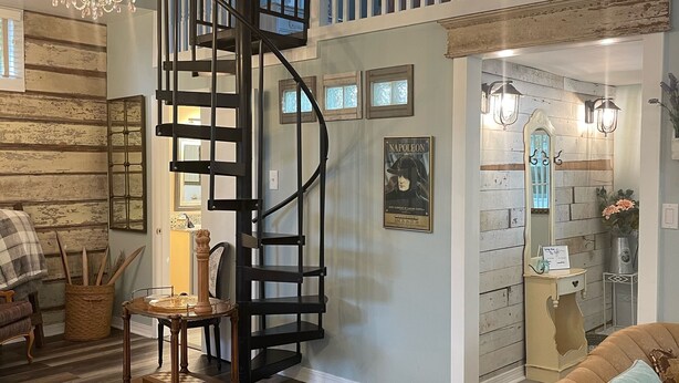 A spacious living area with a wooden floor, a brown sofa, a chandelier, a spiral staircase, and a view of the outdoors through a window. - Adorable Downtown Carriage House Cottage (Natchez)