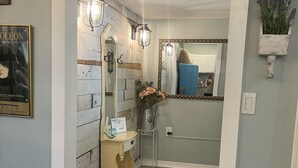 A hallway with a wooden accent wall, a mirror, a small table with a vase of flowers, and a rug on the floor. - Adorable Downtown Carriage House Cottage (Natchez)