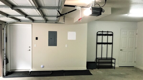 Un garage avec une porte, un luminaire fixé au mur, une étagère en métal noir et un paillasson noir à l’entrée.