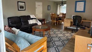 Ein Wohnzimmer mit einem schwarzen Ledersofa, einem Holztisch, einem Esstisch mit Stühlen, einem Schreibtisch mit einem Stuhl und einem gemusterten Teppich.