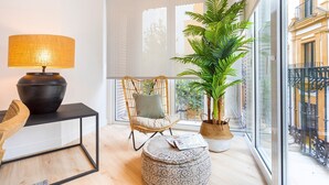 A modern living room with a large window, a wicker chair, a round ottoman, a black table with a lamp, and a potted plant. - Genteel Home Aire II (Sevilla)