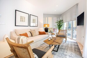 A modern living room with a sofa, coffee table, and wicker chairs. There are framed pictures on the wall, a lamp, and a plant.