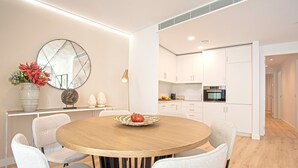 A modern dining area with a round wooden table, white upholstered chairs, a decorative mirror, and a vase with flowers. - Genteel Home Aire II (Sevilla)