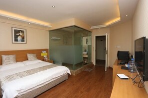 A hotel room with a large bed, a desk with a computer, a television, and a glass partition separating the room from the bathroom.