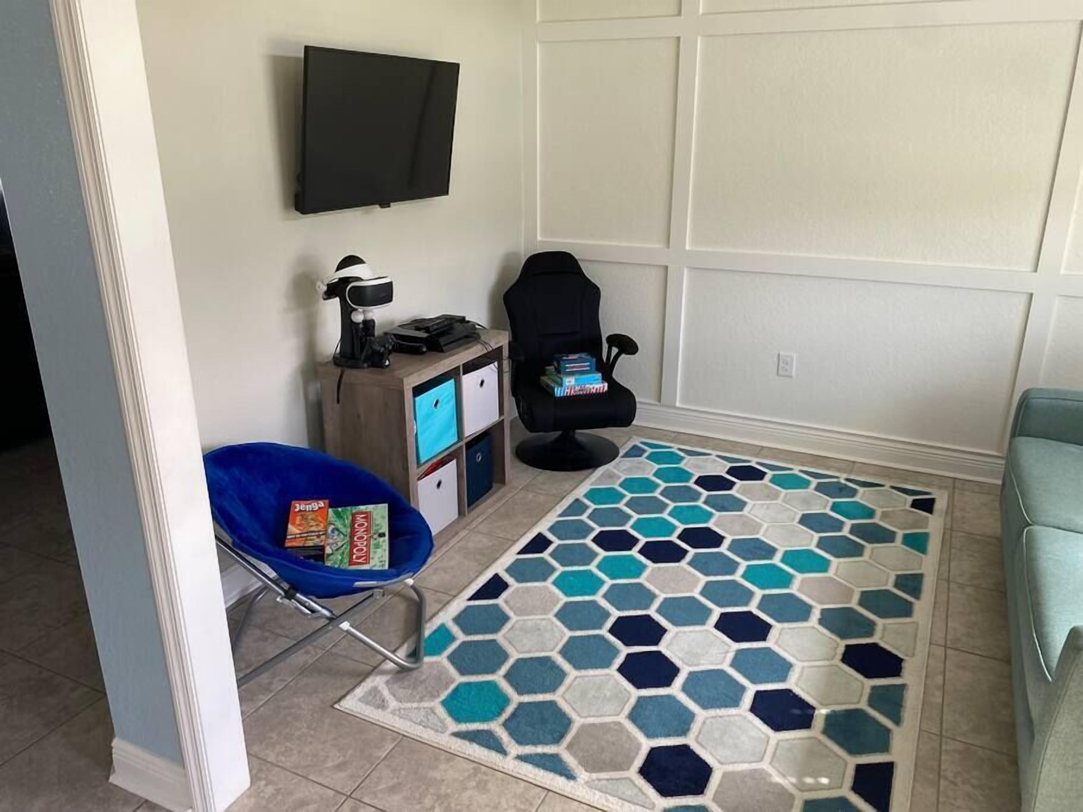 Une pièce avec un téléviseur fixé au mur, un fauteuil de bureau noir, une chaise bleue, un petit meuble en bois avec des rangements et un tapis à motifs.