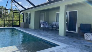 A covered swimming pool area with a glass ceiling, a patio with a table and chairs, and a view of the surrounding greenery. - Gone Coastal! Large heated pool home 30 min from beaches (North Port)