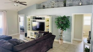 A living room with a dark-colored sofa, a flat-screen TV on a white entertainment unit, a ceiling fan, and a large potted plant. - Gone Coastal! Large heated pool home 30 min from beaches (North Port)