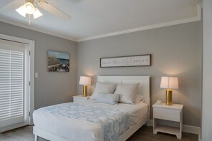 Una camera da letto con un letto, due comodini con lampade, una ventola a soffitto, una citazione incorniciata al muro e una porta con persiane.