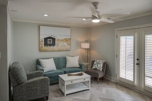Un soggiorno con un divano color petrolio, un tavolino da caffè bianco, una poltrona con motivo decorativo, una lampada da terra e una ventola a soffitto.