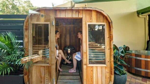Un sauna en bois, avec deux personnes à l’intérieur, une structure extérieure en bois et une vue sur l’extérieur.