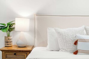A neatly made bed with white linens and decorative pillows, a bedside table with a lamp and a woven vase with a plant, and a plain wall in the background.