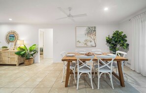 A dining area with a wooden table, white chairs, a ceiling fan, and a beach-themed painting.