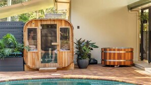 Une structure de sauna en bois dotée de portes en verre, d’un jacuzzi et d’un espace aménagé au bord de la piscine avec des plantes en pot.