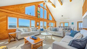 A spacious living area with multiple seating options, large windows, and a high ceiling with wooden beams.