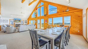 A spacious dining area with a long table set for six, wooden walls, large windows, and a high ceiling with exposed beams.