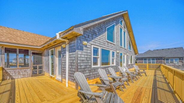 A wooden deck with Adirondack chairs, a house with large windows, and a clear blue sky.