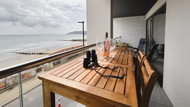 Ein Holztisch mit Weingläsern und einer Flasche, ein Balkon mit Blick auf den Strand und eine Backsteinmauer.