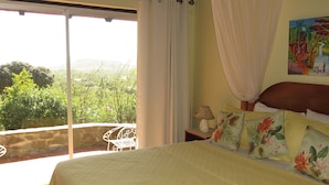 A bedroom with a bed, a nightstand, a lamp, a painting, and a balcony with a view of greenery. - Spanish-style Ocean view Villa set in garden - Calypso Court (Cap Estate)