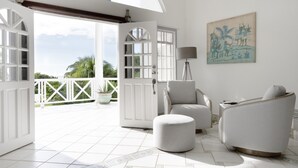 Una habitación amplia con un gran ventanal que da a un balcón, una zona de estar acogedora con una silla y un reposapiés, y un cuadro en la pared.