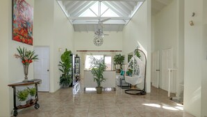 A spacious hallway with a high ceiling, a ceiling fan, and a large window allowing natural light. There are several potted plants and a white hanging chair. - Villa Decaj - Beautiful 2 bedroom Villa with private pool in Cap Estate (Cap Estate)