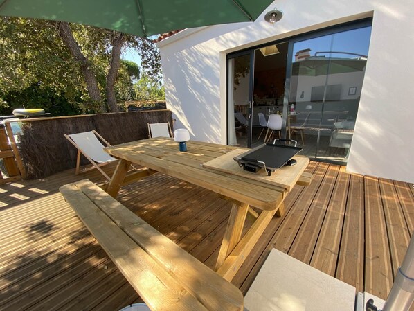 An outdoor wooden deck with a table and chairs, a grill, and a sliding glass door leading to an indoor area.