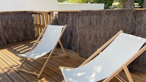 Duas poltronas brancas de tela sobre um deck de madeira, com um muro de palha ao fundo.
