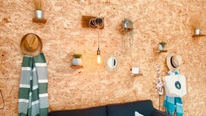 Uma sala de estar acolhedora, com um sofá verde escuro, duas almofadas brancas, uma parede de madeira e diversas decorações penduradas, incluindo um chapéu, um espelho e um lustre.