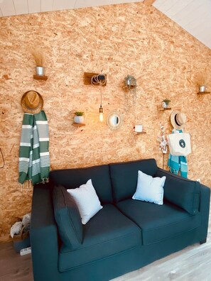 Een gezellige zithoek met een donkergroene bank, twee witte kussens, een houten wand en diverse wanddecoraties, waaronder een hoed, een spiegel en een hanglamp.