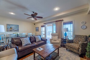 A living room with a brown leather sofa, a patterned armchair, a wooden coffee table, a ceiling fan, and a dining area with white chairs and a table. - Oceanfront 3BR | Indoor & Outdoor Pools + Hot Tub | Pet Friendly | Elevator (Kill Devil Hills)