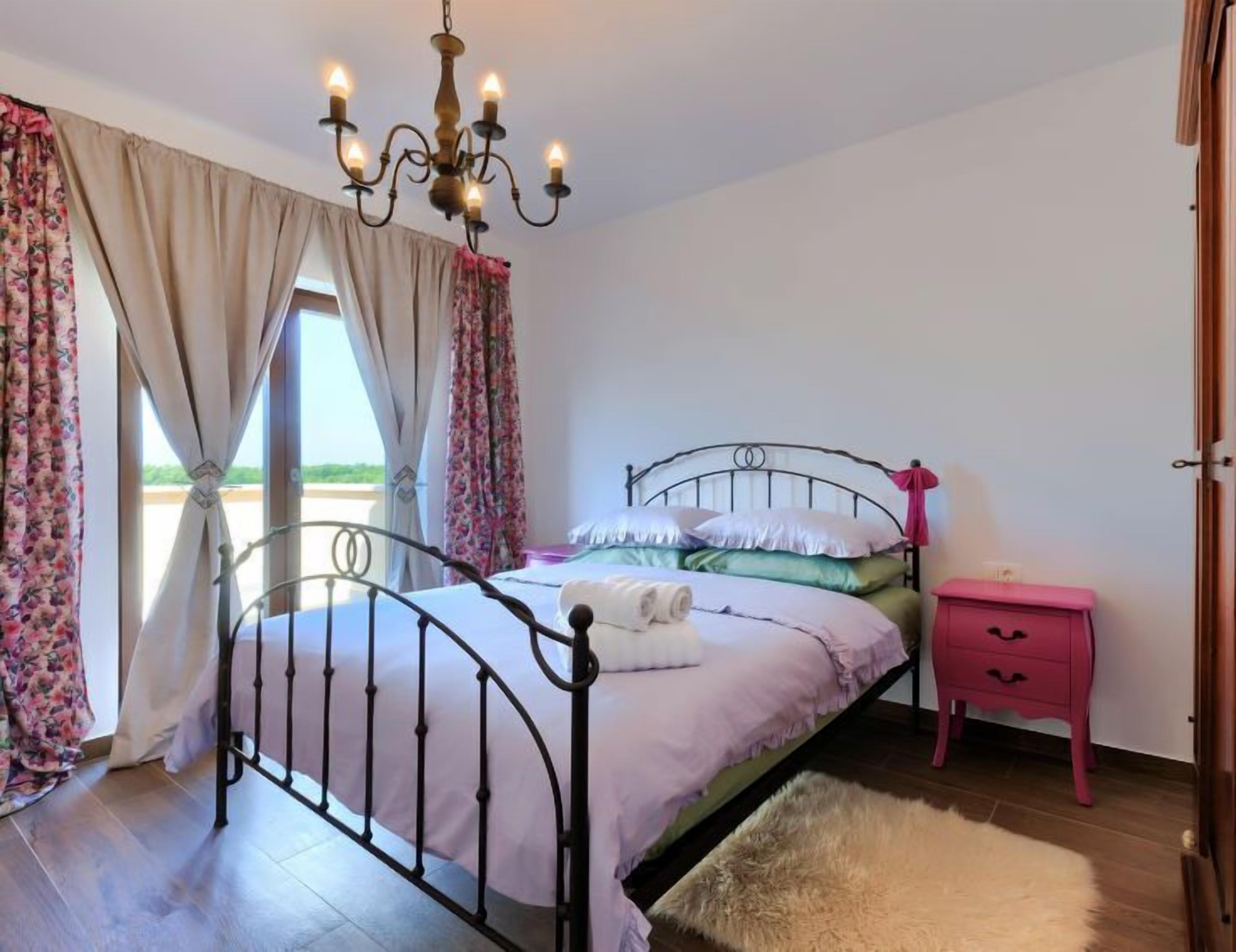 A bedroom with a bed, a nightstand, a dresser, a chandelier, and curtains.