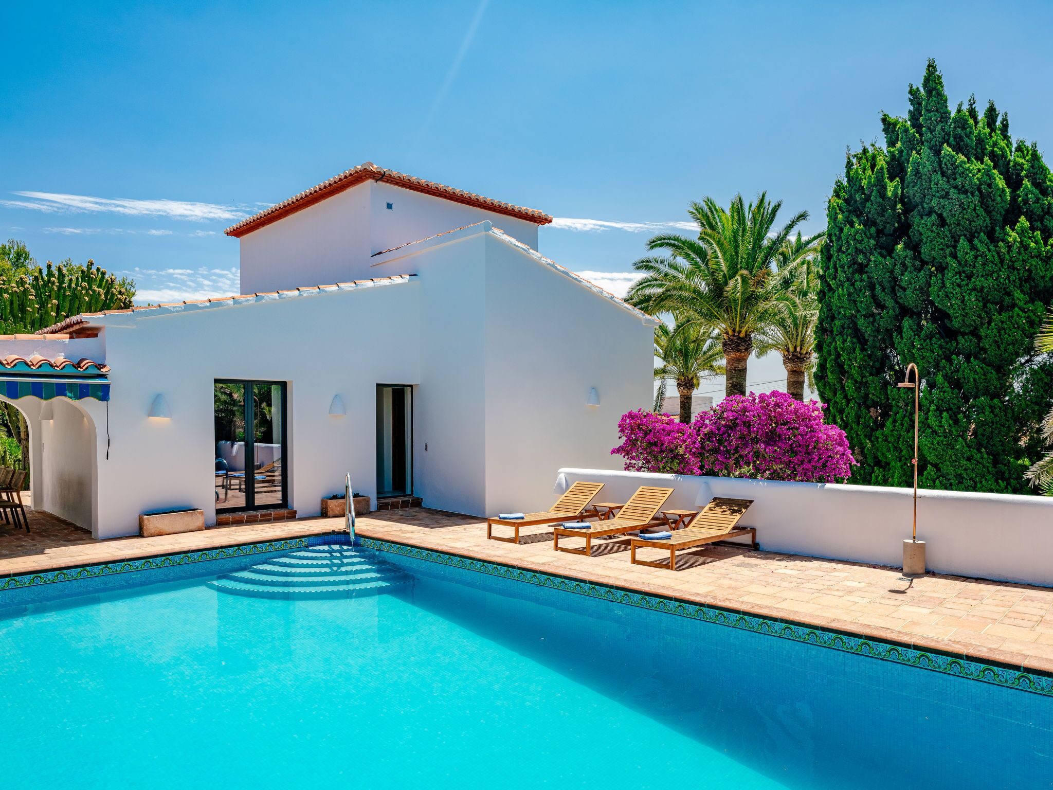 A pool area with a white building, a clear blue pool, and lounge chairs.