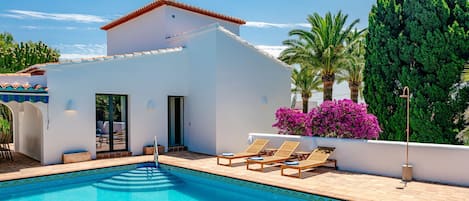 A pool area with a white building, a clear blue pool, and lounge chairs.